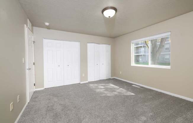 A large empty room with a carpeted floor and a window showing a tree and a building outside.
