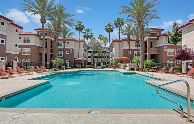 A resort-style swimming pool here at Mozaic at Steele Park surrounded by tall palm trees, comfortable lounge chairs, and shaded patio seating. The sparkling blue water is framed by spacious sundecks and modern apartment buildings, creating a relaxing outdoor retreat perfect for soaking up the Arizona sunshine or enjoying time with neighbors.