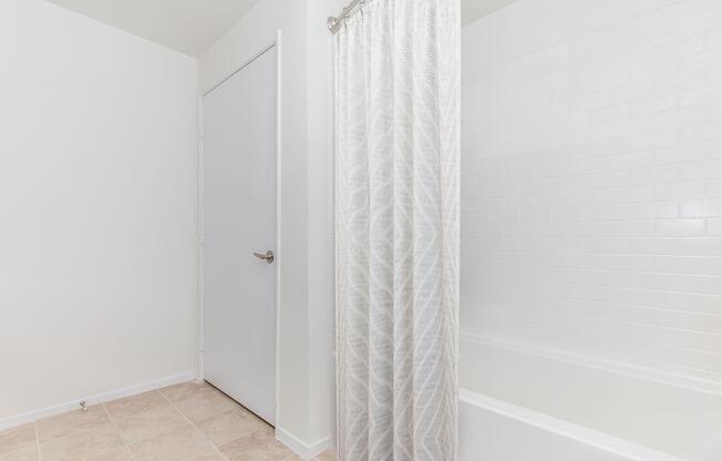 A clean, minimalistic bathroom featuring a white bathtub and a light-colored shower curtain. The walls are painted white, and there is a closed door visible on the left. The floor is tiled in a neutral shade, creating a spacious and airy atmosphere.