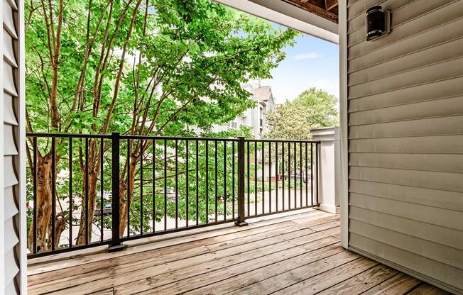 A view of a wooden balcony with a black railing, overlooking a green tree and residential buildings. The sky is clear with a few clouds, creating a bright and inviting atmosphere. The balcony is partially shaded by the overhanging roof above.