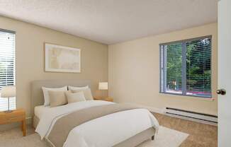 A calm, inviting bedroom here at Adagio featuring plush beige carpeting, soft neutral walls, and a comfortable bed with layered white and neutral bedding, complemented by matching wood bedside tables and lamps, while large windows with blinds allow natural light and offer peaceful greenery views outside.