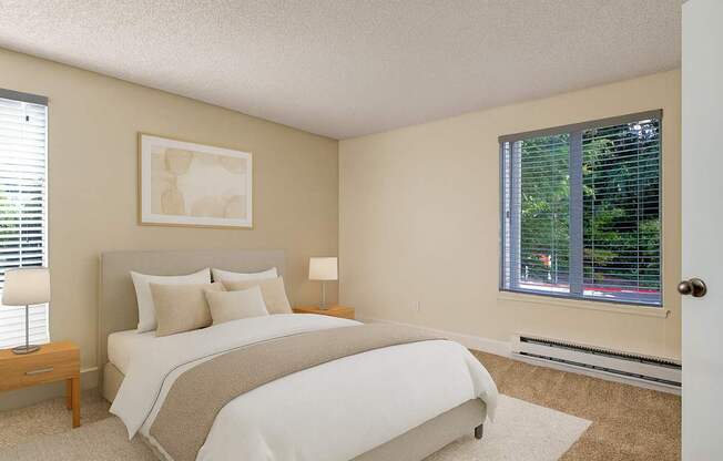 A calm, inviting bedroom here at Adagio featuring plush beige carpeting, soft neutral walls, and a comfortable bed with layered white and neutral bedding, complemented by matching wood bedside tables and lamps, while large windows with blinds allow natural light and offer peaceful greenery views outside.