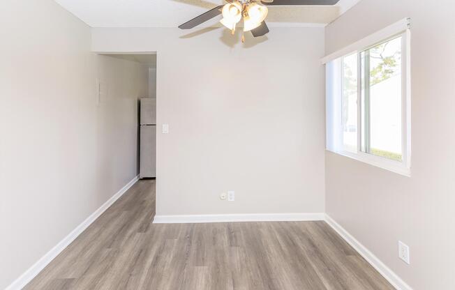 A spacious, empty room featuring a ceiling fan, light-colored walls, and modern laminate flooring. Natural light enters through a window on one side, while a doorway leads to a small kitchenette area visible in the background. The overall atmosphere is bright and inviting.