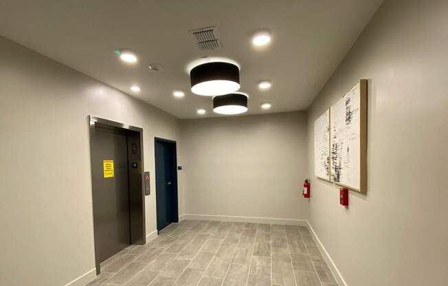 A hallway leading to a waiting area with elevators, wall art, tile-style flooring, and modern circular lights.