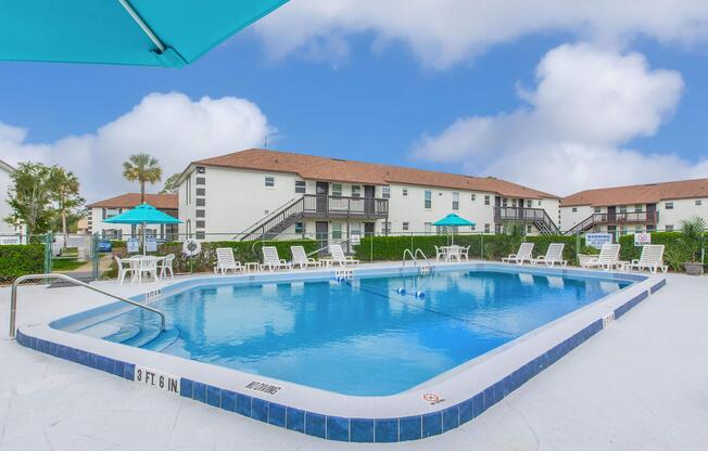 A clear blue swimming pool surrounded by white lounge chairs and umbrellas, set against a backdrop of two-story residential buildings. The area is landscaped with green bushes and palm trees, under a bright blue sky with scattered clouds. The pool depth is marked as 3 to 6 feet.