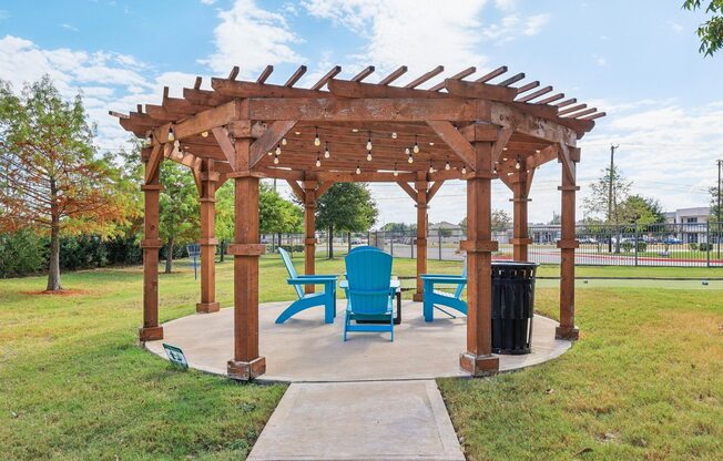 A wooden pergola with blue chairs and a trash can is in a grassy area.