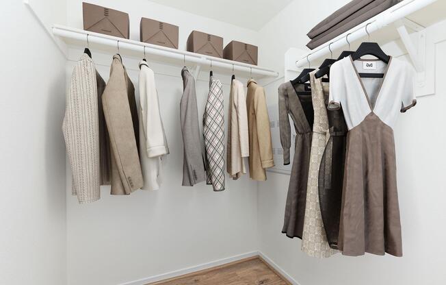 A neatly organized closet featuring a variety of clothing items hanging on a rack. The garments are in neutral tones, including dresses and blouses, with several boxes on a shelf above. The closet has a clean, minimalist design with a light-colored wall and wooden flooring.