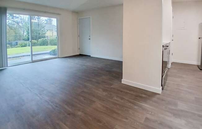 A spacious room with wooden flooring and a view of a green outdoor area through the windows.