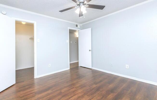 A spacious, empty room with light gray walls and a ceiling fan. Wooden laminate flooring stretches across the floor. Two white doors are visible: one leading to a closet and another to a nearby room, both slightly ajar. The overall atmosphere is bright and inviting, with ample natural light.