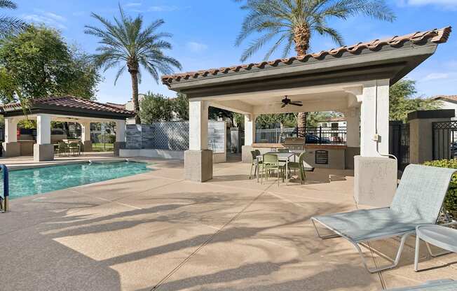 A patio with a table and chairs is surrounded by a pool and palm trees.