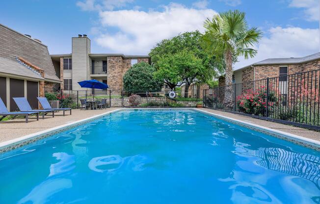 A clear, blue swimming pool surrounded by lounge chairs, with lush greenery and flowering bushes in the background. There are buildings in the distance and a blue umbrella providing shade near the pool area.