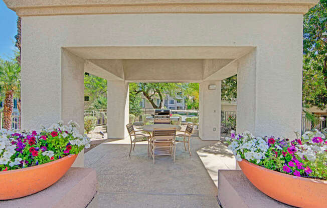 A shaded outdoor ramada here at Mirador featuring a covered concrete patio with a round dining table and chairs, surrounded by vibrant flower planters in large terracotta pots. A built-in grill and prep area sit just beyond the seating, creating a welcoming space for outdoor dining and relaxed gatherings among mature trees and landscaped grounds.