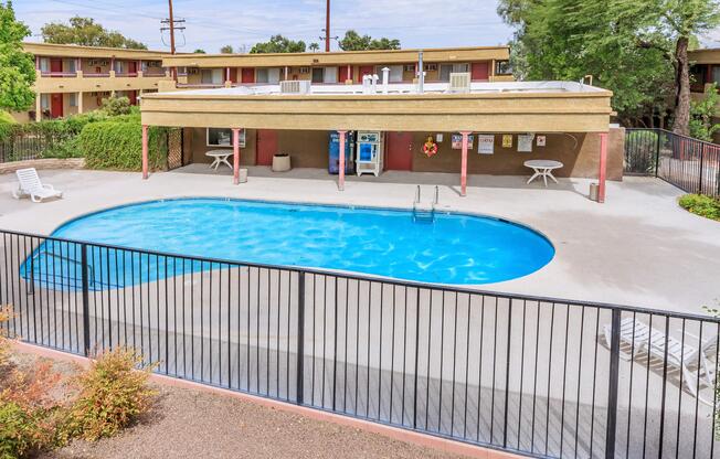 A clean, inviting swimming pool surrounded by a fence. The pool area features several lounge chairs and a shaded seating area with a table. In the background, there are two-story buildings with balconies. Lush greenery surrounds the pool, creating a relaxing atmosphere.
