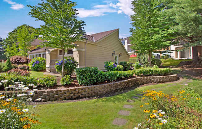 A beautifully landscaped community setting here at Adagio featuring manicured lawns, colorful flower beds, and mature trees surrounding a charming clubhouse building, with stone retaining walls and winding walking paths that create a peaceful, park-like atmosphere ideal for strolling, relaxing outdoors, and enjoying the natural surroundings.