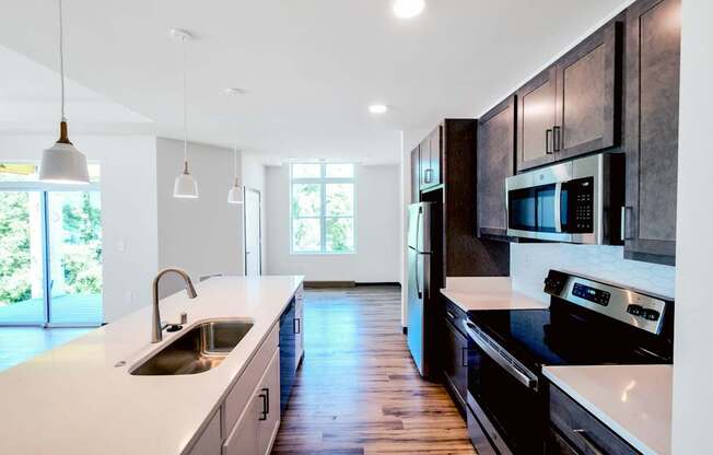 A modern kitchen here at Van Buren East with stainless steel appliances and dark wood cabinets. A long center island with sink and pendant lighting overlooks an open living area with access to a private balcony.