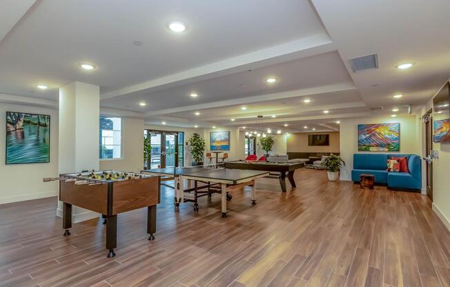 A spacious game room featuring a foosball table, a billiards table, and a vibrant lounge area with a blue couch. The room is well-lit with recessed lighting and has large windows, showcasing colorful artwork on the walls and plants for decoration.