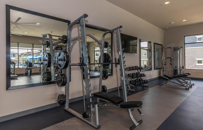 A modern gym interior featuring a weightlifting machine with plates, a bench press, free weights on a rack, and an exercise bench. Large mirrors and windows enhance the space, providing ample natural light and a motivating environment for workouts.