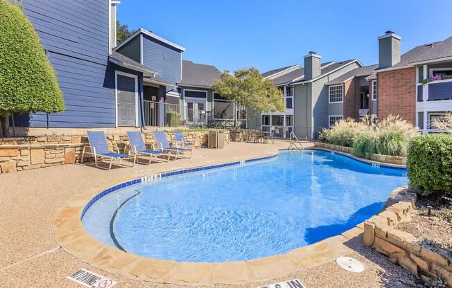 A well-maintained outdoor swimming pool surrounded by lounge chairs, with a vibrant landscape of shrubs and grass. The setting features modern apartment buildings in the background under a clear blue sky. The pool has a gentle curve and a depth marker indicating a maximum depth of 6 feet.