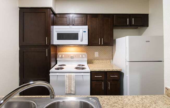 A kitchen with dark wood cabinets and white appliances in a 1 bedroom, 1 bath B, 629 sq ft apartment home at The Biltmore Apartments located in the Vickery Midtown neighborhood of Dallas, TX.