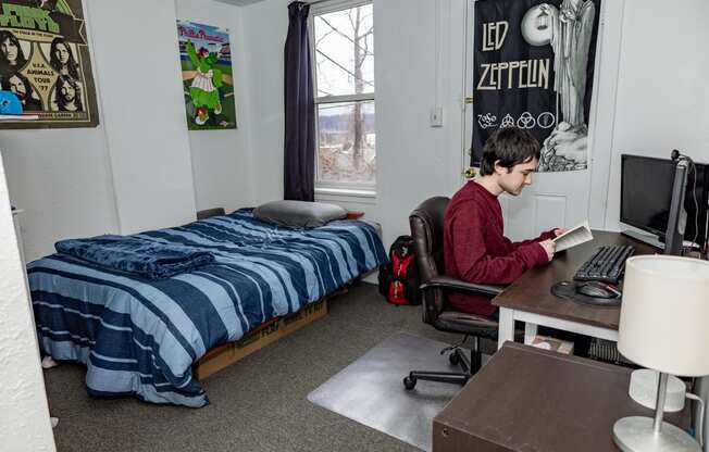 a student sits at a desk in a dorm room with a bed in the background