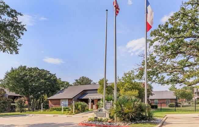 A brick building with a sloped roof, surrounded by trees and landscaping. In front, two flags are raised on flagpoles. A sign is displayed in the foreground, and the area appears well-maintained with a clear blue sky overhead.