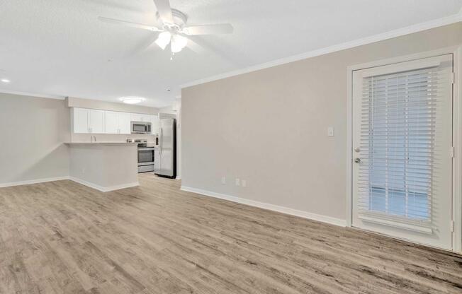 Interior of a modern apartment featuring an open floor plan. The living area has light wood-like flooring, a ceiling fan, and a doorway with blinds leading outside. The kitchen, visible in the background, has white cabinets and stainless steel appliances.