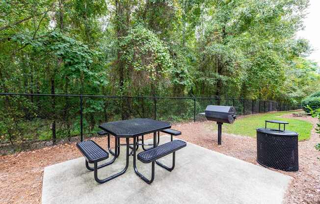 A black metal table with four benches is surrounded by a fence and trees.