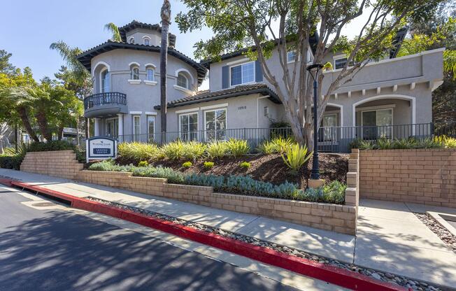 A modern two-story house with a unique architectural design, featuring a round tower, balconies, and large windows. The front yard is landscaped with plants and trees, and the property has a sign that reads "California Resort Management." The street in front has red curb markings.