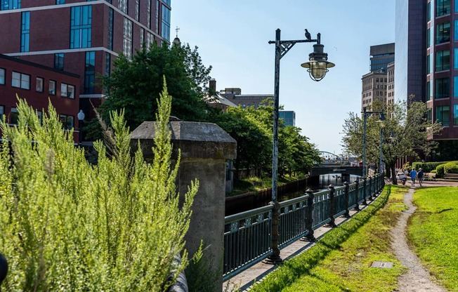 a path next to a river with tall buildings in the background