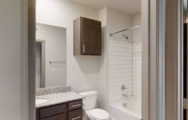 Modern bathroom here at 49 West Apartments with white tile bathtub and shower, dark wood cabinets, granite countertop, large wall mirror, and neutral walls. Clean and minimalistic.