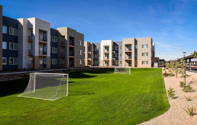 A soccer goal is in the foreground of a grassy area in front of apartment buildings.