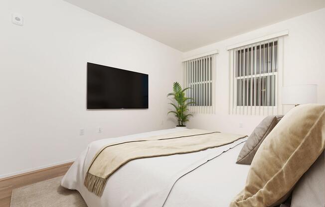 A modern bedroom featuring a neatly made bed with beige and gray pillows, a light-colored throw blanket, a large flat-screen TV mounted on the wall, and two windows with white blinds. A potted plant adds a touch of greenery to the space, enhancing the contemporary aesthetic.