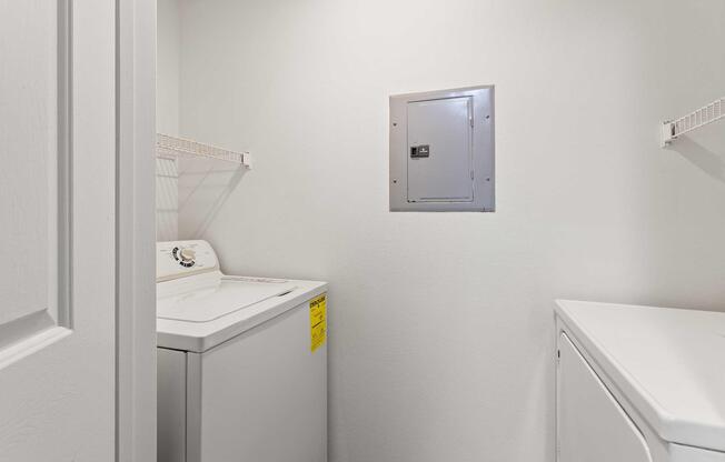 A laundry room featuring a washing machine and a dryer side by side. There is a utility panel on the wall and metal shelves above the appliances for storage. The walls are painted white, creating a clean and simple appearance.