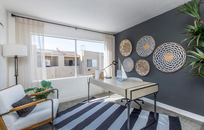 A modern home office featuring a wooden desk with a lamp, a stylish armchair, and decorative wall hangings. Large windows allow natural light to flow in, and a potted plant adds a touch of greenery. The room has a neutral color palette with a dark accent wall and a striped area rug.
