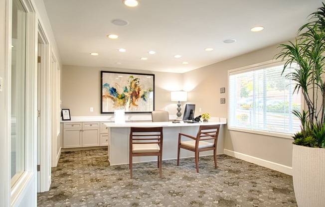 A welcoming leasing office here at Andorra featuring a bright reception desk with two guest chairs, built-in cabinetry with a countertop workspace, neutral carpeting with a subtle pattern, recessed ceiling lighting, large windows with blinds bringing in natural light, abstract wall art, and a potted plant that adds a polished, professional feel.