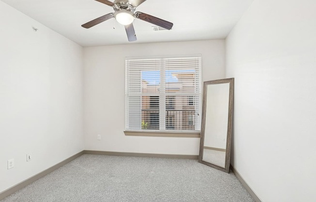 A bedroom with a ceiling fan, mirror, and a window with blinds at GreenVue Apartments in Richardson, TX