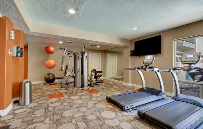 A fitness room here at Bridges with treadmills, a cable weight machine, wall mirrors, mounted TV, patterned carpet, and orange accent walls with exercise balls and strength equipment.