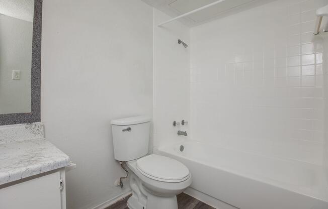 A clean, modern bathroom featuring a white bathtub with a showerhead, a white toilet, and a simple countertop with a sink. The walls are painted in light colors, and the floor has a wood-like finish, creating a bright and inviting space.