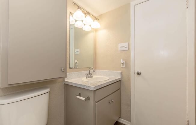 A clean, modern bathroom featuring a light-colored countertop with a sink, mirrored light fixture above, gray cabinetry for storage, and a white toilet. The walls are painted a soft beige, and a closed door is visible on the right side of the image.