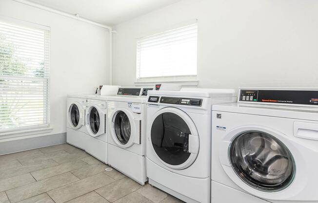 A bright laundry room featuring four modern white washing machines lined up against a wall. The room has light-colored tile flooring and natural light coming in through a window with blinds.