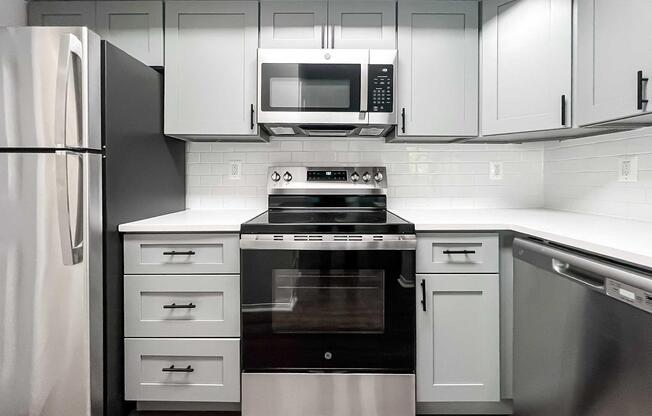 Modern kitchen featuring light gray cabinets, stainless steel appliances including a refrigerator, microwave, and oven. The countertop is white, and there's a dishwasher next to the oven. The backsplash consists of white tiles, creating a clean and contemporary look.
