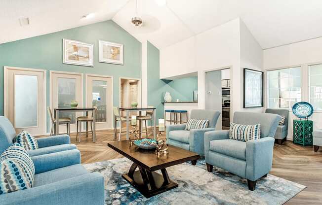 Clubhouse interior with wood style flooring, accent rug, coffee table, high top tables and chairs, vaulted ceiling and blue accents with kitchen in background