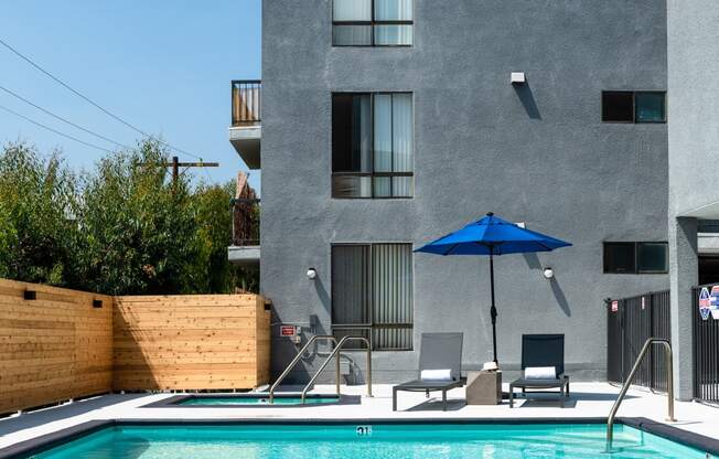 a pool with a blue umbrella and two chairs in front of a gray building