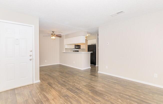 A spacious, unfurnished living room featuring light-colored walls, laminate flooring, and a ceiling fan. There's an entrance on the left and an open kitchen area visible in the background, complete with a breakfast bar. The overall ambiance is bright and inviting, suitable for personal decoration.