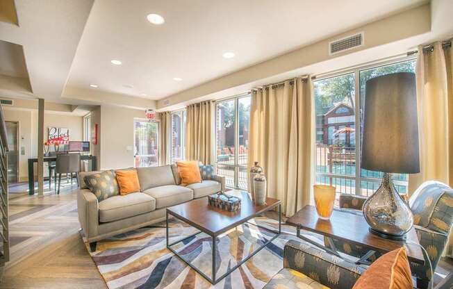 A bright clubhouse lounge here at Lakewood on the Trail with a gray sofa, patterned accent chairs, wood coffee table, large windows, and pool views, creating a warm, welcoming feel.
