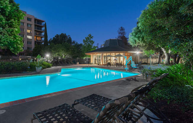 Palo Alto, California Apartments - Tan Plaza - Gated Pool Area at Night, with Lush Greenery, Lounge Chair, and White Umbrellas