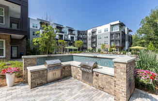 an outdoor kitchen with two grills next to a pool with an apartment building in the background at Century University City, Charlotte, NC