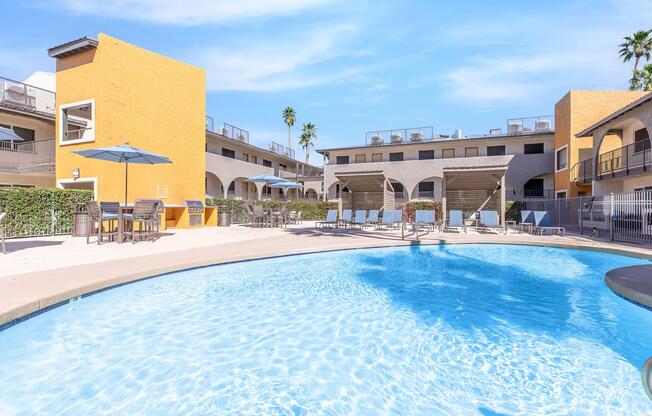 A bright and inviting swimming pool surrounded by lounge chairs and tables, with palm trees in the background. The pool area is located near colorful buildings featuring a sunny yellow exterior, creating a relaxing atmosphere for outdoor lounging and leisure.