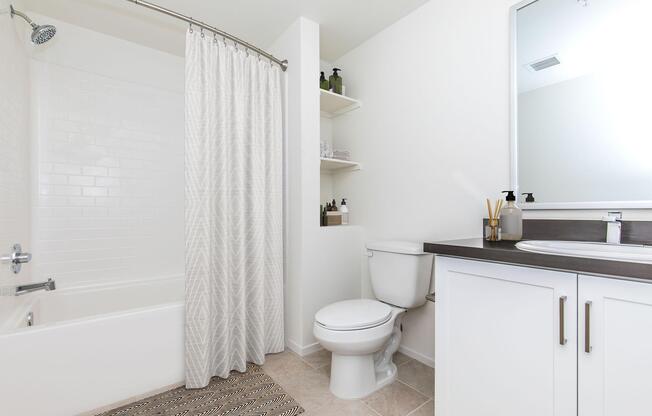 A clean and modern bathroom featuring a bathtub with a shower curtain, a toilet, and a sleek vanity with a sink. The walls are painted white, and there are shelves with neatly arranged toiletries. A textured rug is placed on the tiled floor, adding warmth to the space. Natural light brightens the room.