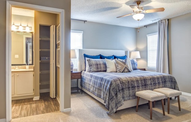 Staged bedroom with two windows, gray walls, carpet, bed with seating in front, two nightstands with lamps, ceiling fan, and entry way to en-suite bathroom in background.